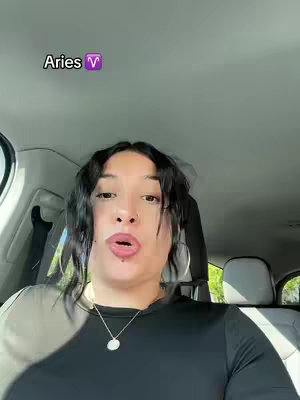 A selfie-style shot taken inside a car. The main subject, a woman with dark hair, is positioned in the center of the frame, looking towards the camera with an open mouth. She is wearing a black long-sleeved shirt and a necklace. Behind her is the interior of the car, visible through the windshield, and the top of the seat is visible. The background is slightly blurred, emphasizing the subject. Text overlay with Aries.