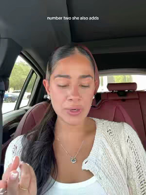 A woman with dark hair and a white sweater is seated in a car, likely filming a video. The background shows the interior of the car, including the headrest of the other seat. Overhead the roof of the car is visible. A text overlay at the top says "number two she also adds".