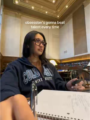 A woman wearing glasses and a Columbia University hoodie is seated in a library, looking off-screen, with a notebook and pen on a desk in front of her. Above her, text overlays read, "obsession's gonna beat talent every time."