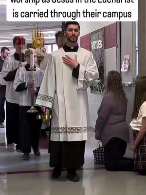 A man in white religious vestments leads a procession through a hallway. Other figures, some in similar outfits, follow. On the right, two people kneel in prayer. The image is well-lit, with a warm color palette.