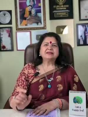 A woman in a patterned blouse with dark hair and a bindi is seated in a chair. She is in an office space with framed pictures hanging on the wall behind her. A sign reading, "I am a Happy Soul" is visible on a nearby table. A microphone is attached to her blouse.