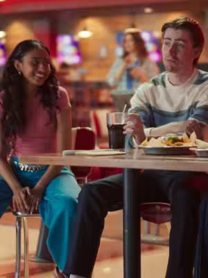 A couple and a friend in a diner setting. The girl is the main character in the picture looking towards the guy. She has a pink top and her friend is sitting with her. The other party is sitting across of them. The background is a bit blurry.