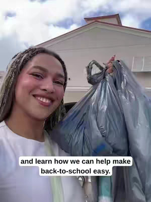 A smiling person holds multiple blue shopping bags in front of a house. The person has their hair in braids and is wearing light makeup. The background includes a white building with a decorative roof. Text at the bottom of the image reads: "and learn how we can help make back-to-school easy."