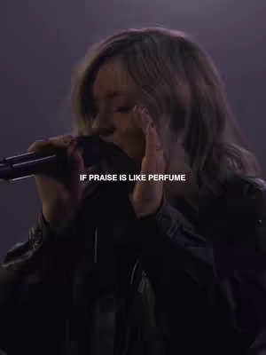 A woman with grey hair, facing slightly left, singing into a microphone. She is dressed in black clothing with some of her upper body visible. The backdrop is dark, with soft lighting creating a hazy atmosphere. Text overlayed on image reads: IF PRAISE IS LIKE PERFUME