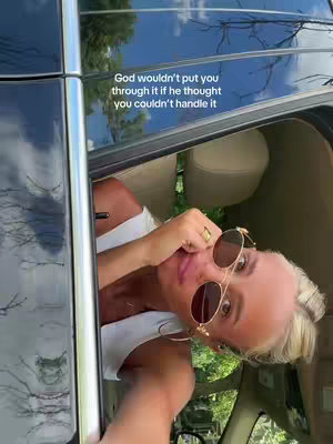 The image shows a woman in the front seat of a car, with her head and shoulders visible. She's looking towards the camera with sunglasses on and a ring on her finger. The car window is open, and there's text overlayed at the top. The background shows the interior and exterior of the car with nature peeking through.