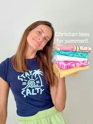 A woman with long brown hair stands in front of a light gray background. She's wearing a navy blue t-shirt printed with a white design that says "Stay Salty". She's holding a stack of colorful t-shirts. The tops are mostly pink, white, and teal, and there is text overlayed "Christian tees for summer!!"