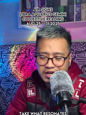 A man with glasses and grey hair is in front of a microphone, the background has shelves and a rainbow colored light.