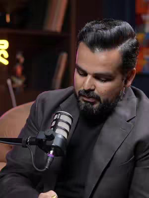 A man with a beard sits at a desk holding a microphone. He's wearing a dark gray suit jacket over a black shirt. The background suggests a recording studio or office setting with bookshelves.