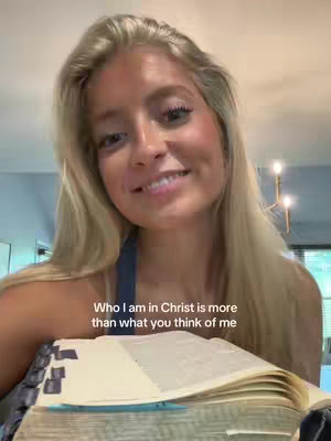 A young woman with long blonde hair smiles at the camera while holding an open book. The background is slightly blurred, suggesting an indoor setting. Text overlay mentions the woman in Christ.