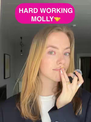 A woman with blonde hair is holding a finger to her lips, with text overlay in a pink background. She is wearing a blazer over a white shirt. The background is an interior setting.
