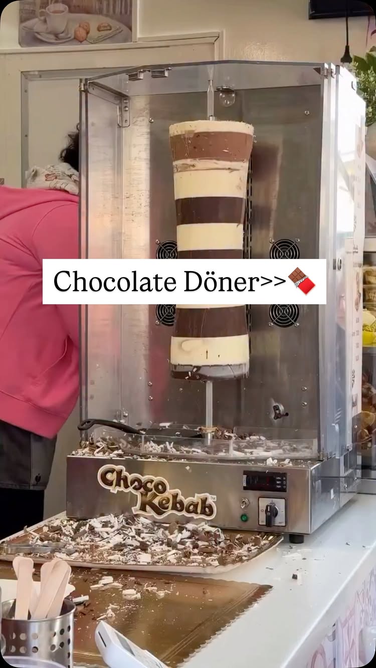 The cover image shows a machine with a rotating cylinder of layered chocolate, resembling a döner kebab. A white text box with the words "Chocolate Döner" is overlaid with a chocolate bar emoji. A person wearing a pink hoodie can be seen in the background blurred, behind the machine.