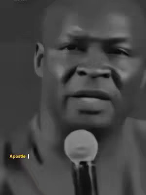 A black and white close-up shot featuring a Black man speaking into a microphone. The image is focused on his face, with a soft spotlight highlighting the central focus of the scene. The background is a dark, nondescript space that accentuates the speaker's presence. Positioned directly in front of him, a prominent microphone stands out, suggesting a moment of public speaking or recording. The text 'Apostle |' is placed near the left side of the lower portion of the frame.