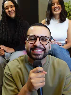 The cover image shows a man centered in the foreground with glasses, a slight beard, and a microphone, smiling towards the camera. Behind him, two women are seated on either side, slightly out of focus, also smiling. The background appears to be a blurred interior setting with a neutral color palette, contributing to the overall warmth of the image
