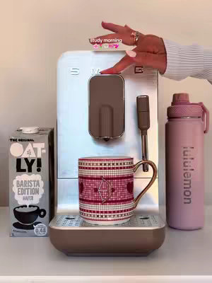A person's hand is pressing the button on a coffee machine. To the left is a carton of Oatly oat milk. To the right is a pink lululemon water bottle. In front is a coffee mug.