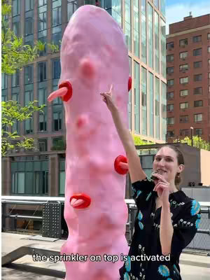 A vertical shot captures an outdoor scene with a modern city backdrop. Dominating the frame is a large, abstract sculpture resembling a pink, elongated foot with red accents. A woman stands to the right of the sculpture, extending her arm as if pointing at it. She smiles slightly, wearing a dark floral patterned top. Behind them, there are tall buildings and green trees under a bright sky.