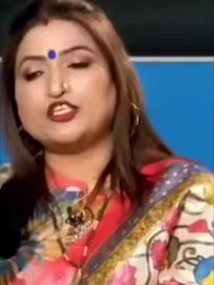 A woman with brown hair, a red bindi on her forehead and a colorful traditional style sari is looking and talking into the camera. The image has a close up face with slightly blurred background.