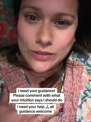 A selfie-style video shows a woman from the chest up, with a soft, possibly indoor, lighting. She looks directly at the camera with a concerned expression. The background is blurred, but appears to be a mix of floral and dark colors. The woman has light skin, dark hair and is wearing a top that has a floral pattern mixed with a black fabric. Several lines of white text are overlaid on the image at the center of the frame, addressing the viewer and asking for intuitive guidance.