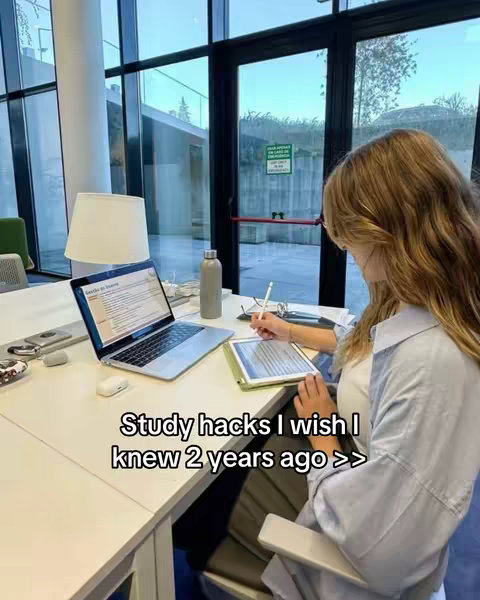 A person sits at a desk in a modern, well-lit space, likely a study or library. They are working on a laptop and a tablet, holding a stylus and pen. The image is framed from an angle, with the focus on the person's workstation.