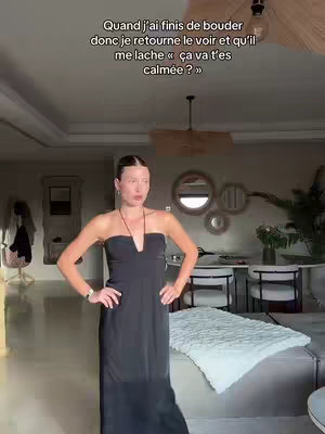 A woman in a black dress stands indoors with her arms crossed, exuding confidence. Text in French is overlaid at the top, and the background includes a living room setting with mirrors and furniture.