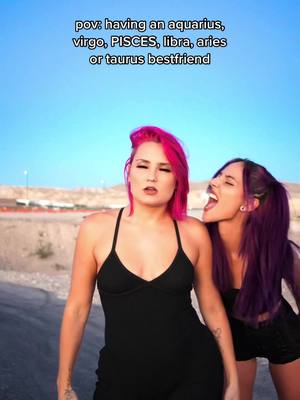 Two women are captured in a shot, one with vibrant pink hair and a black dress and the other with purple hair and a matching outfit. The pink-haired woman has a look of bemusement, while her companion playfully seems to bite her. The background shows a road and a landscape under a clear blue sky.
