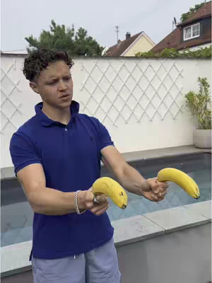 A man with curly hair in a blue polo shirt is holding two yellow bananas like guns, looking to the left of the frame with a slightly concerned expression. His left wrist has a silver bracelet. He is standing outside near a pool and a white fence.