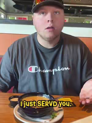 A young man with fair skin, a short haircut, and a slight smile is captured in a medium shot from the chest up. He is wearing a dark gray t-shirt with the red and white Champion logo prominently displayed on the front. He is also wearing a black baseball cap. His eyes are open wide, and his expression suggests surprise or shock. He's seated at a restaurant, with a bowl of soup and a small dipping sauce in front of him. The table is made of dark wood.