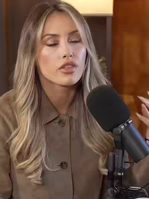A woman with long blonde hair is centered in the frame, facing slightly upward with her eyes closed. She wears a tan button-down shirt, and a microphone is positioned in front of her. The background is blurred, suggesting an indoor setting.