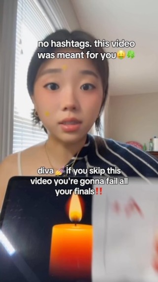 A young woman with fair skin and dark hair is positioned in a medium shot, looking directly at the camera with a gentle expression. She has light makeup, including blush and a dab of yellow color. Text overlays are positioned at the top and bottom of the frame; a lit candle is visible in the bottom. The background is a mix of indoor space and blurred outdoor elements.