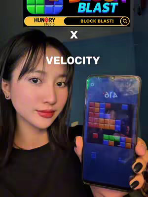 A young woman with long dark hair and red lipstick holds a smartphone displaying the Block Blast game in front of her. The image is set in a room with a blue wall in the background. Text overlays the image, including game branding and the word 'VELOCITY'.