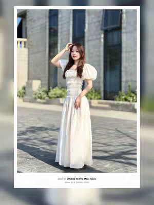 A woman in a white dress poses outdoors, with a modern building in the background. She is holding her hand up to her head, and there is text in the bottom left. The image is cropped in a white border.