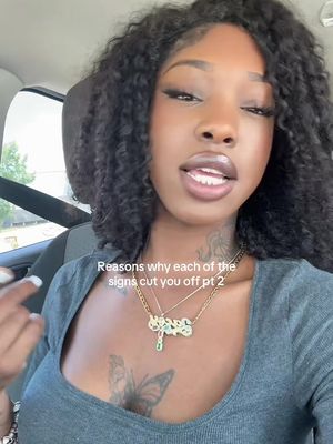 A woman with long, dark, curly hair and full lips is shown in a selfie-style shot from inside a car. She has visible makeup, a necklace, and a tattoo. The background is a car interior with a blurred view.
