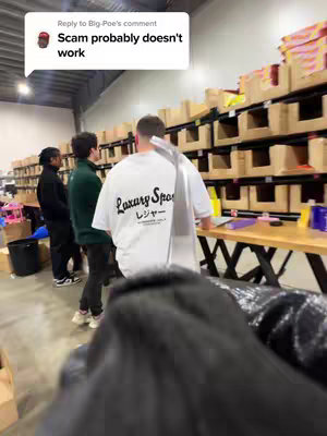 The image shows a warehouse setting. Three men are visible: a man with his back to the camera wearing a white t-shirt with black printed text, another wearing a green shirt and the third in black attire. They are positioned in front of shelves filled with various boxes. A speech bubble from a comment is in the top left with the text: "Scam probably doesn't work ".