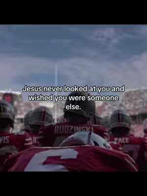 The image shows the backs of football players in red uniforms on a field. Text overlaid on the image reads, 'Jesus never looked at you and wished you were someone else.' The sky is overcast, and the atmosphere is somber.