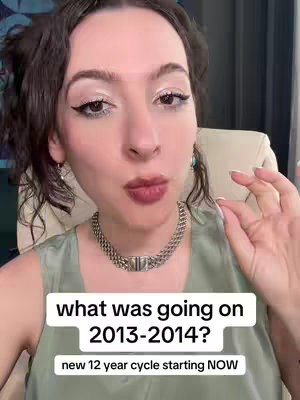 A selfie-style shot shows a woman with dark wavy hair and professionally applied makeup, facing the camera. A text overlay is positioned below her, with a background that highlights the text and includes the question "what was going on 2013-2014?" and, just below, "new 12 year cycle starting NOW". Her choice of attire includes a silver necklace and a light green-colored blouse. The background shows a neutral, light-colored setting behind her head and shoulders.