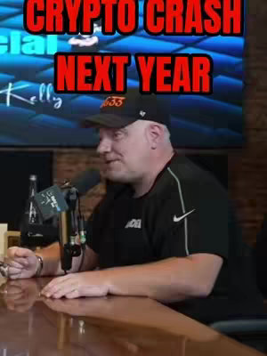 A man is seated, speaking into a microphone, with a blurred background featuring a TV screen. The text overlay reads "CRYPTO CRASH NEXT YEAR" in large red letters, positioned above the man's head. The man is wearing a black shirt and a black cap, with "33" on front.