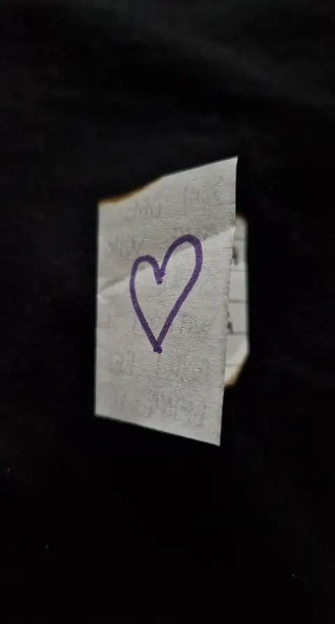 A piece of paper folded into a small, irregular shape with a purple heart drawn on its visible surface. The paper sits against a black background, creating a contrast that highlights the heart.