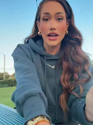 A woman with long brown hair and fair skin, wearing a blue Nike hoodie and gold hoop earrings, is shown from the chest up. She is looking at the camera, with her mouth slightly open as if speaking. The background shows a bit of blue sky and green vegetation. The composition is a medium close-up, putting emphasis on her face. The lighting appears natural.