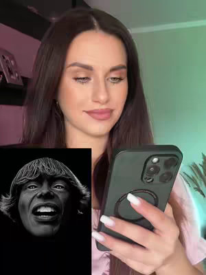 A woman with long brown hair, wearing a pink top holds a smartphone, displaying an image of a black and white drawing of a man's face with an open mouth. The room is lit with shades of green and pink. The overall composition highlights both the woman and the phone screen, focusing on the contrast between the artwork and the woman's gaze.