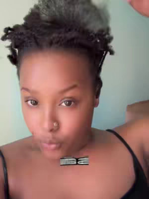 Close-up shot of a person with dark hair styled in small, twisted sections on top of their head. They are looking directly at the camera with a neutral expression, slightly pursing their lips. They have a nose ring and dark eyes. The background is a solid, light teal color. The person is wearing a black tank top.