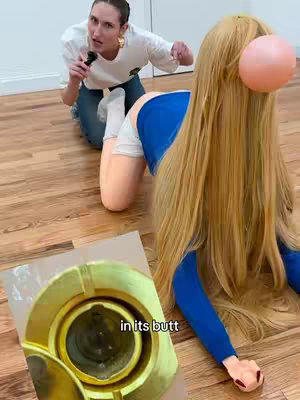 A woman kneels on a wooden floor, holding a microphone and looking towards a doll-like figure that is positioned on its hands and knees. The figure, wearing a blue top, white shorts, and a wig, has a pink balloon attached to its head. A golden object with an opening is in the foreground.