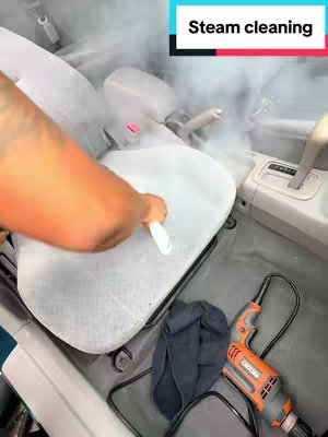 The image shows the interior of a car with a person steam cleaning the driver's seat. The seat is light gray and a steam cleaner is being used on the surface. Steam is visible rising from the seat. There is a black cloth and an orange and black steam cleaner on the floor of the car. The text overlay indicates the video is about steam cleaning.