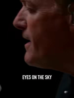 A close-up shot featuring the side profile of a man's face, set against a black background. The focus is on his face, with his mouth slightly open as if he is speaking or singing. The lighting accentuates the contours of his face, making the details of his skin visible. The text overlayed on the image in white, which reads "EYES ON THE SKY".
