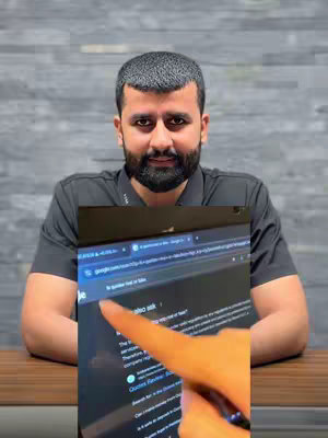 A man with a beard is seated at a table, with a laptop positioned in front of him. He is pointing at the screen with his right index finger. The background is a light gray wall. The laptop screen displays a webpage with text and a search bar that shows 'google.com'.