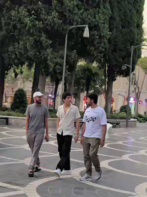Three men are walking in a paved area with trees and streetlights in the background. The man in the middle is wearing a white shirt and black pants. The other men are wearing t-shirts and casual pants. The sky is overcast.
