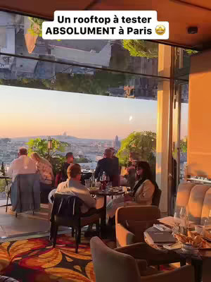 A photo of a rooftop restaurant at golden hour, with a view of Paris and several people seated at tables. The foreground has tables set with glasses and drinks, and a floral pattern on the floor. The background shows people sitting at tables on a balcony with a view of rooftops and buildings in Paris. A text overlay is present at the top, and the overall scene is warm and inviting, bathed in the soft light of sunset.