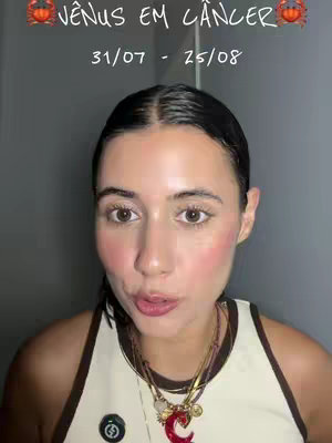A selfie-style close-up with a woman as the central figure. She’s looking directly at the camera with a subtle smile, light skin, and a natural makeup look with prominent blush. She wears a light brown tank top with dark brown trim, and a collection of layered necklaces with various pendants and charms are visible around her neck. Above her is a text overlay with zodiac and astrological signs. The background is shadowed, creating a focus on the creator.