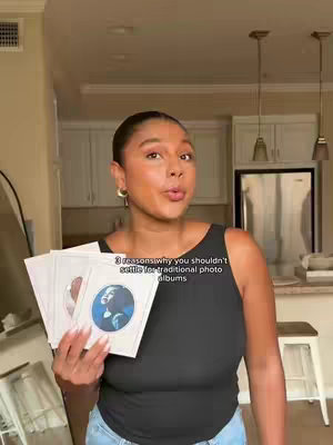 A woman with dark hair and a black tank top holds a stack of white rectangular cards, displaying a portrait image on the bottom card. The background includes a home interior with white cabinets and a glimpse of a stainless steel refrigerator.
