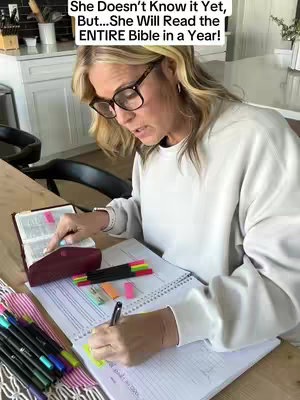 A woman wearing glasses is sitting at a wooden table, reading a Bible with colorful pens, highlighters, and a notebook. The background shows a kitchen.