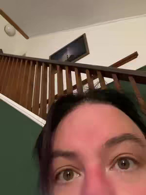 A close-up shot focuses on a person's eyes and a portion of their forehead, with a staircase and a picture frame visible in the background.