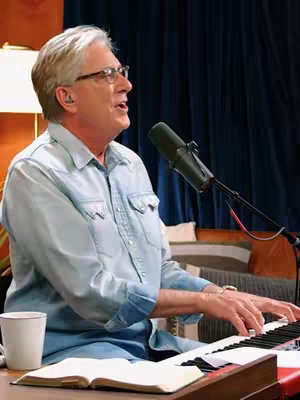 A man plays a piano in a warmly lit setting. He wears glasses and a blue shirt. A microphone is positioned in front of him. To the side is a coffee cup, and an open book sits on the piano.